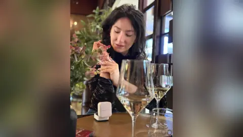 Woman in a black outfit sitting at a restaurant table with a glass of white wine in a cozy atmosphere.