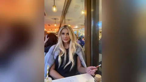 Blonde woman in a grey faux fur coat sitting at a restaurant table with a stylish, elegant mood.