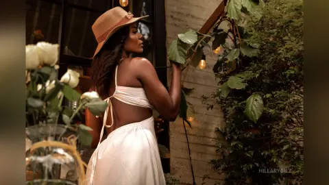 Elegant woman in a white sundress and straw hat standing by lush green plants in a warm, sunlit garden setting.