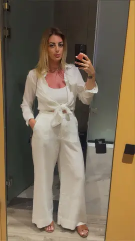 Woman in white linen two-piece outfit taking a mirror selfie in a modern bathroom setting.