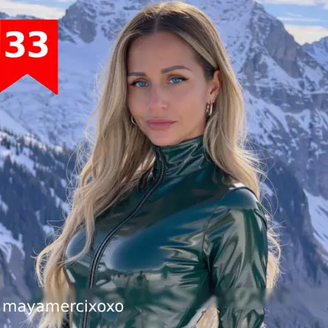 Blonde woman in a dark green shiny turtleneck outfit posing against a snowy mountain landscape.