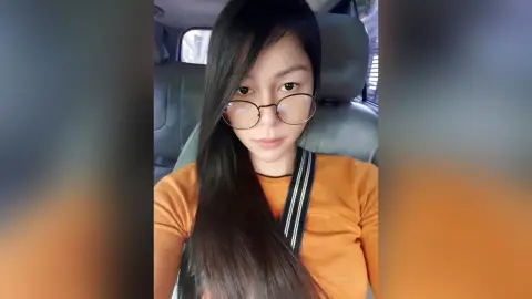 Young woman with long dark hair and glasses wearing an orange top sitting in a car interior.