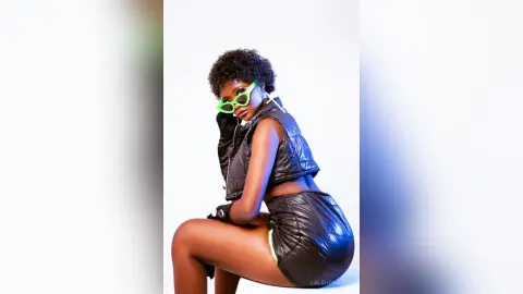 Stylish Black woman in black leather outfit and green sunglasses posing confidently against a white background.