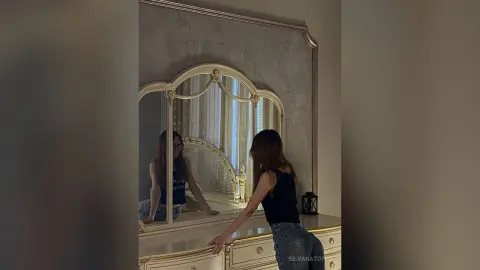 Young woman in a black tank top and jeans looking into an ornate vintage gold mirror in a moody room.