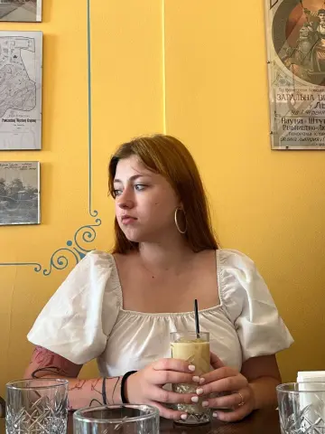 Young woman in a white puff-sleeve blouse sitting at a cafe table with a cold drink, thoughtful mood.