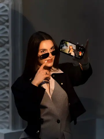 Stylish woman in a black blazer and grey waistcoat taking a mirror selfie with sunglasses in soft lighting.