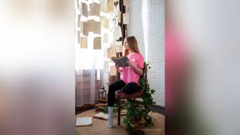 AuroraCruise sits on a chair reading a book, wearing a pink t-shirt and black leggings in a bright room with white brick walls and hanging paper decorations.