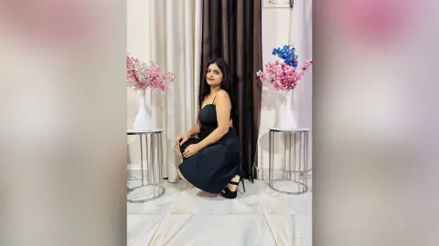 Elegant woman in a black cocktail dress sitting in a bright room with pink floral arrangements.