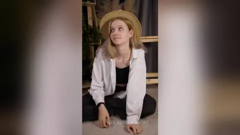 Smiling young woman in a white button-down shirt and straw hat posing in a cozy indoor setting.