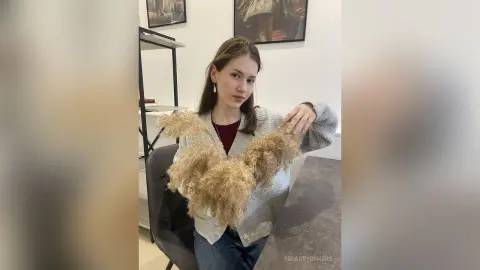 Young woman in a grey cardigan holding a large bouquet of dried pampas grass in a bright modern room.
