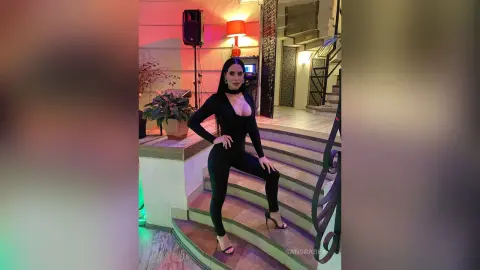 Elegant woman in a sleek black bodysuit posing on indoor stairs near a warm lamp and potted plants.