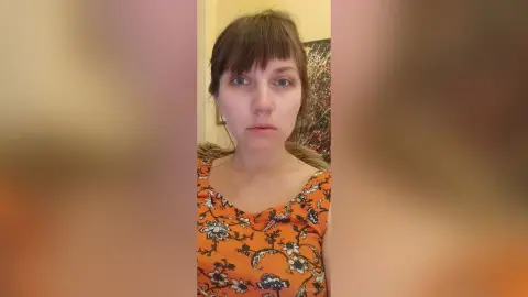 StacyMalvin looks at the camera with brown hair and bangs, wearing an orange floral print tank top.