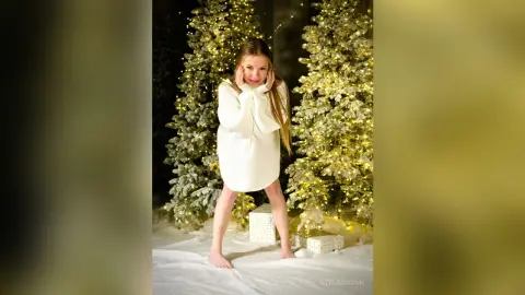 StelaBrown wears a white knit sweater dress while standing between two lit Christmas trees.