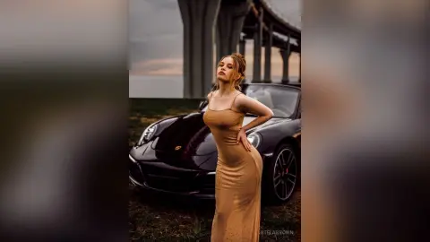 StelaBrown poses outdoors in a tight, tan sleeveless dress that hugs her curves, standing next to a black car under a bridge.