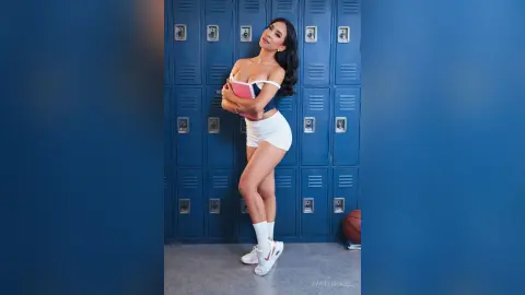 Smiling woman in a white crop top and skirt posing in front of blue school lockers.