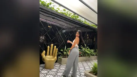 Smiling woman in white crop top and grey wide-leg trousers posing in a modern indoor garden setting.
