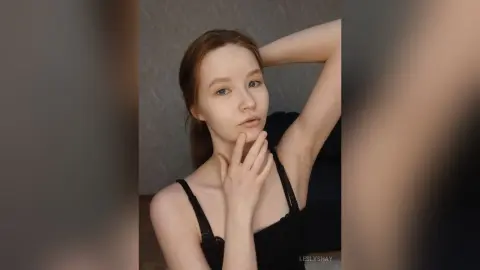 Young woman in a black camisole tank top posing thoughtfully against a neutral gray background.