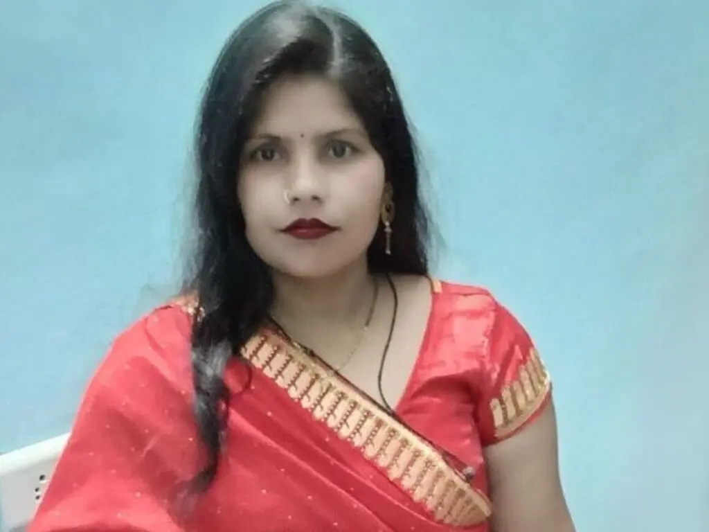 SaronLey wears a red and gold patterned saree, posing against a light blue background with her long black hair draped over her shoulder.