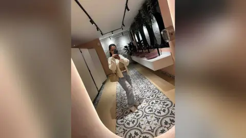 Young woman in a white blouse and light denim jeans taking a mirror selfie in a modern minimalist hallway.