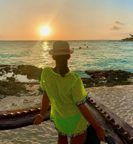 misssLush — misssLush, wearing a bright neon yellow mesh shirt and a white hat, looks out at the ocean during a sunset on a beach.