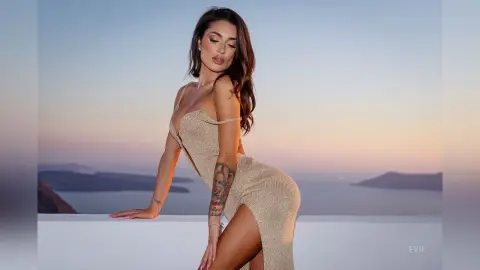 Elegant woman in a shimmering gold evening gown posing against a sunset seascape in Santorini.