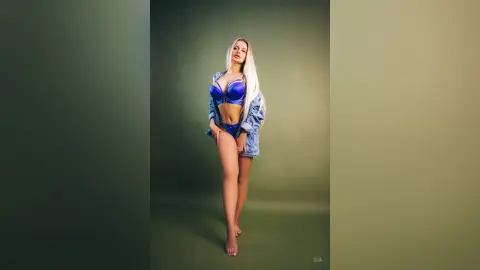 Gia stands against a green background wearing a blue satin bra and matching blue panties with an open denim jacket.