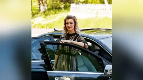 Elegant woman in a black lace dress leaning against a luxury car in a sunny garden setting.