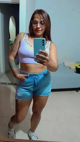 Woman in white crop top and denim shorts taking a mirror selfie in a casual indoor setting.