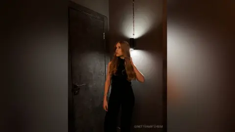 Moody portrait of a blonde woman in a black turtleneck standing by a door in dim, dramatic lighting.