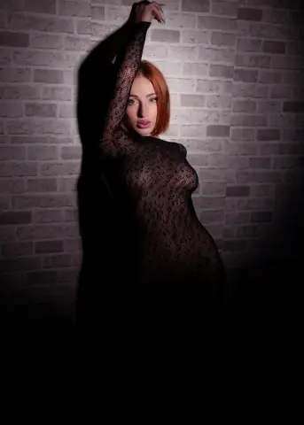 Redheaded woman in a black lace bodysuit posing against a rustic brick wall in moody lighting.