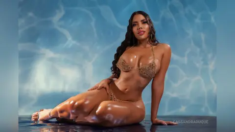 CassandraDuque poses in a gold lace bikini against a blue water-patterned background, showing off her wet, glowing skin and long dark hair.