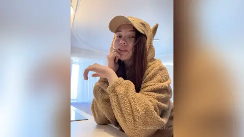 Young woman in a cozy tan fleece hoodie and baseball cap relaxing in a bright, airy room.