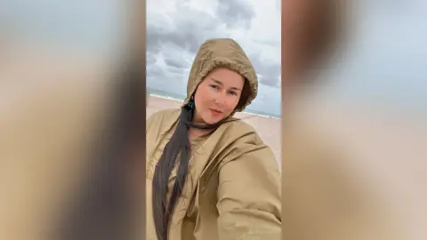 Smiling woman in a tan hooded raincoat standing on a cloudy beach.