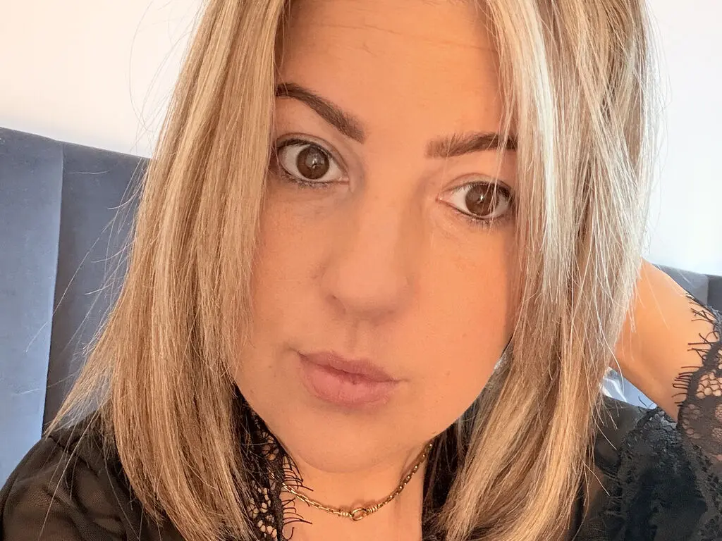 BelleWarick looks directly at the camera with her blonde hair framing her face, wearing a black lace top and a gold necklace.