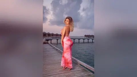 Blonde woman in a pink maxi dress standing on a wooden pier at sunset by tropical ocean villas.