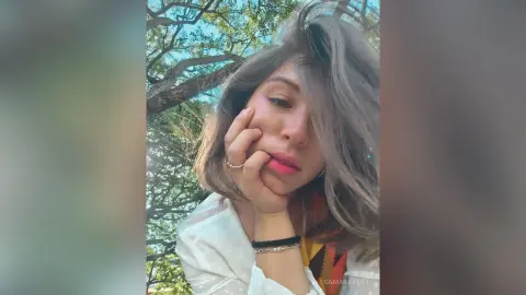 SamaraBert poses with her hand against her cheek, showing her long light-brown hair and pink lipstick against a background of green trees.