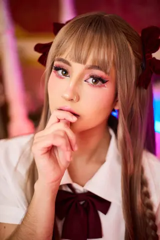 Young woman with blonde bangs and pink eye makeup wearing a white shirt and burgundy bow in soft lighting.