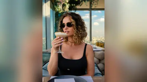 Stylish woman in a black tank top sipping a cocktail at a sunny rooftop lounge.