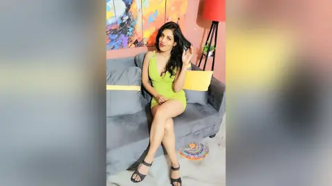 TaraLeo wearing a short lime green dress, sitting on a grey couch and running her hand through her dark hair.