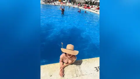 Smiling woman in a straw hat and bikini relaxing by a bright blue resort swimming pool on a sunny summer day.