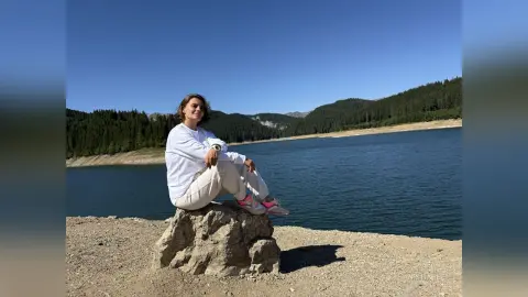 Young woman in white sweatshirt sitting on a rock by a scenic mountain lake under a clear blue sky.
