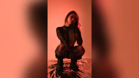 RosieThomas — RosieThomas poses in a black lace bodysuit and black boots against a red background, squatting low on a shiny, reflective surface.