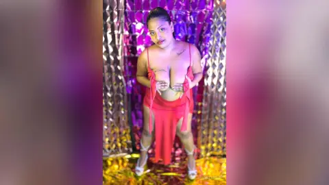 Curvy woman in a red mini dress posing against a shimmering gold sequin backdrop in moody pink lighting.