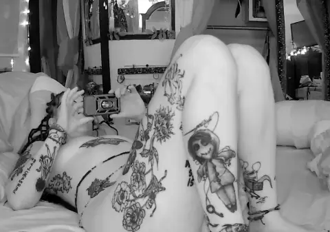 Black and white photo of a tattooed woman lying on a bed holding a vintage camera in a cozy bedroom setting.