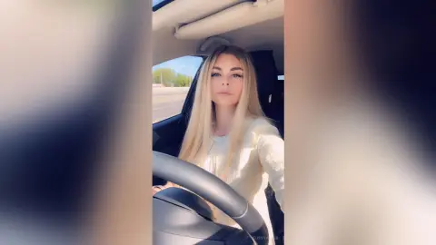 Blonde woman in a white sweater sitting in a car on a sunny day with a calm expression.