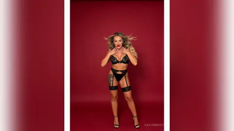 KaylinWhite poses in black lace lingerie and stockings against a red background, running her hands through her blonde hair.