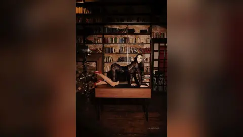 Elegant woman in a black bodysuit posing on a desk in a moody, vintage library setting.