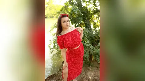 Young woman in a red polka dot summer outfit posing by a peaceful river in natural sunlight.