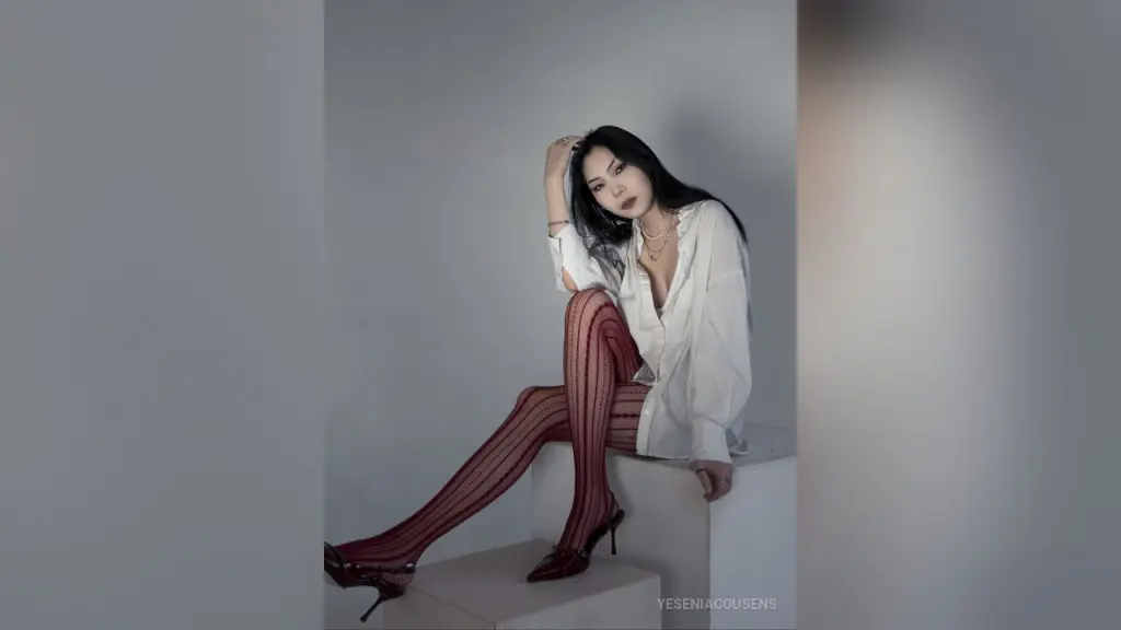Elegant woman with long black hair in a white button-down shirt posing against a grey studio background.
