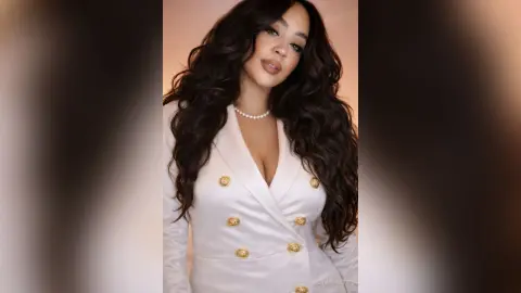 Glamorous woman with wavy dark hair wearing a white satin blazer and pearl necklace in a soft studio setting.